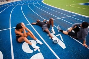 Three athletes rest on a blue track, exhausted after a run. Two sit, one lies down, all catching their breath