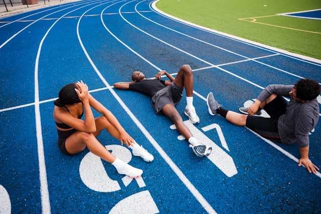 Three athletes rest on a blue track, exhausted after a run. Two sit, one lies down, all catching their breath